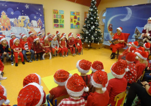 Grupa dzieci siedzi na krzesełkach wokół pani dyrektor Marii Królikowskiej, która czyta legendę o świętym Mikołaju, w tle dekoracja świąteczna, choinki.